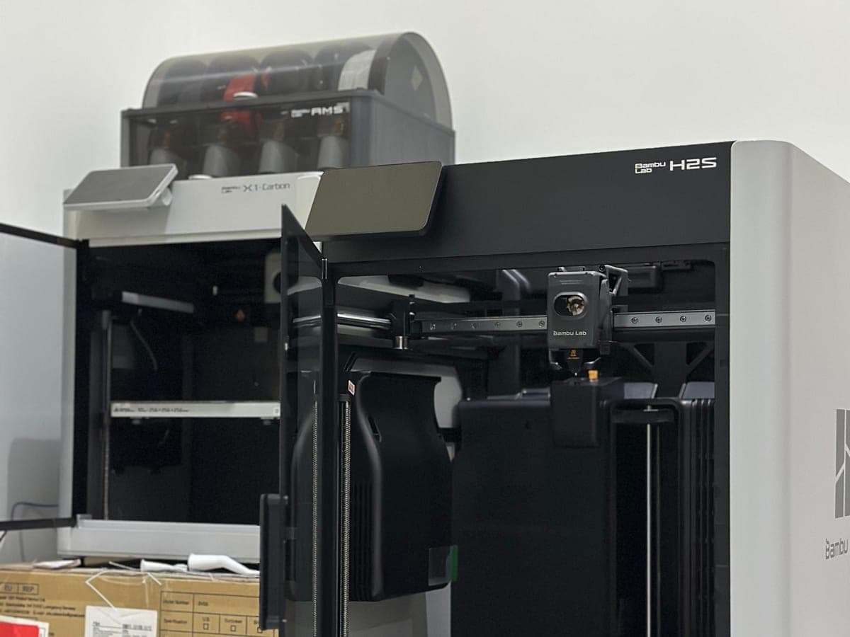 A Bambu Lab H2S standing open beside an X1 Carbon with its filament changer on top